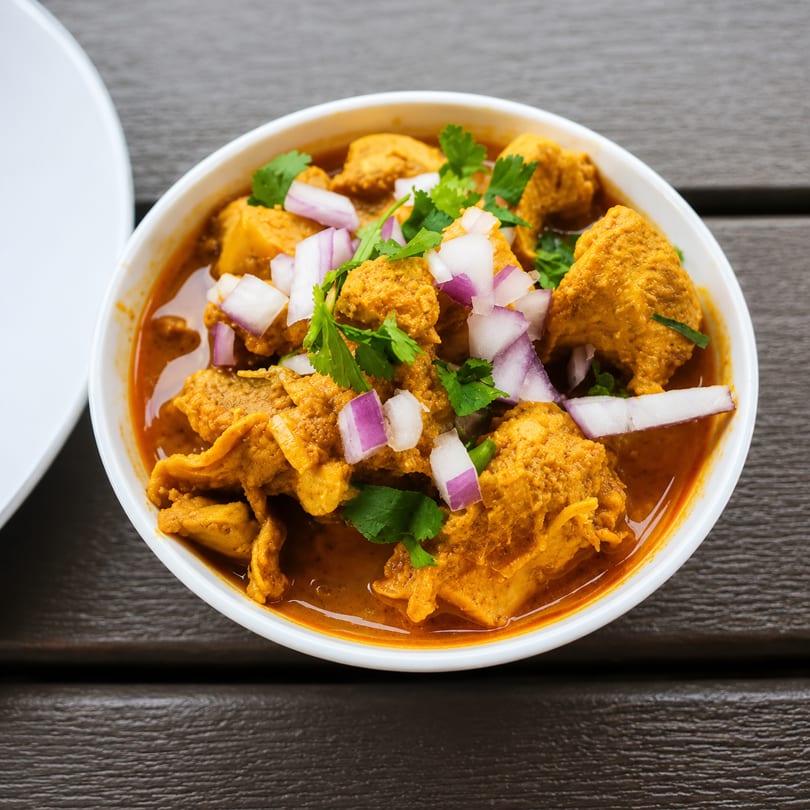 A bowl of chicken curry with red onions and cilantro on a dark wooden table.