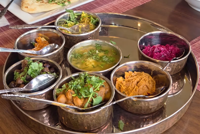 A silver platter holds seven small bowls of various Indian dishes, including curries, soup, and a red relish.
