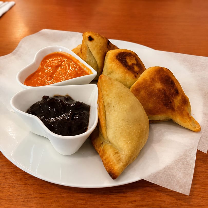 Golden-brown empanadas with two dipping sauces, one orange and one dark, on a white plate.