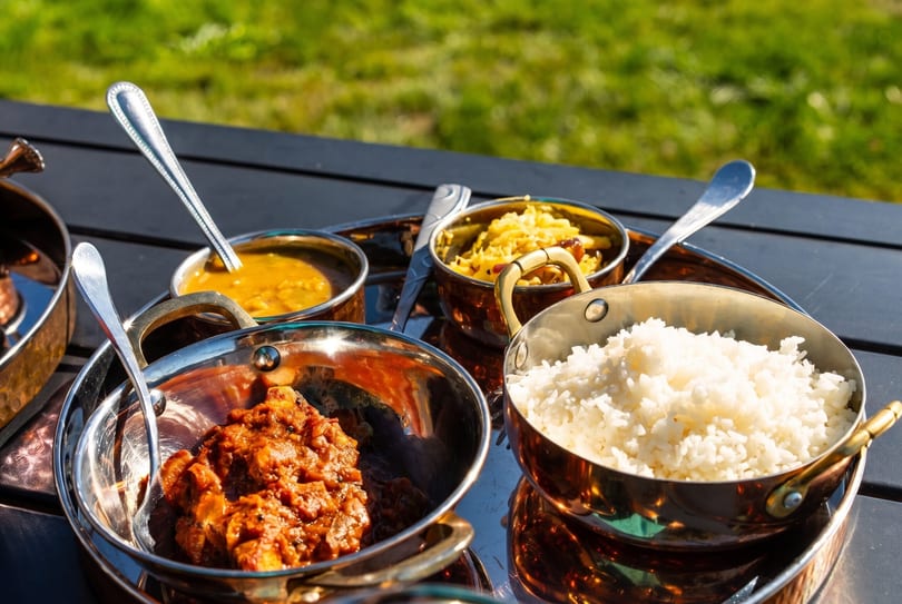 Indian thali with rice, curry, and other dishes in metal bowls on a dark table outdoors.
